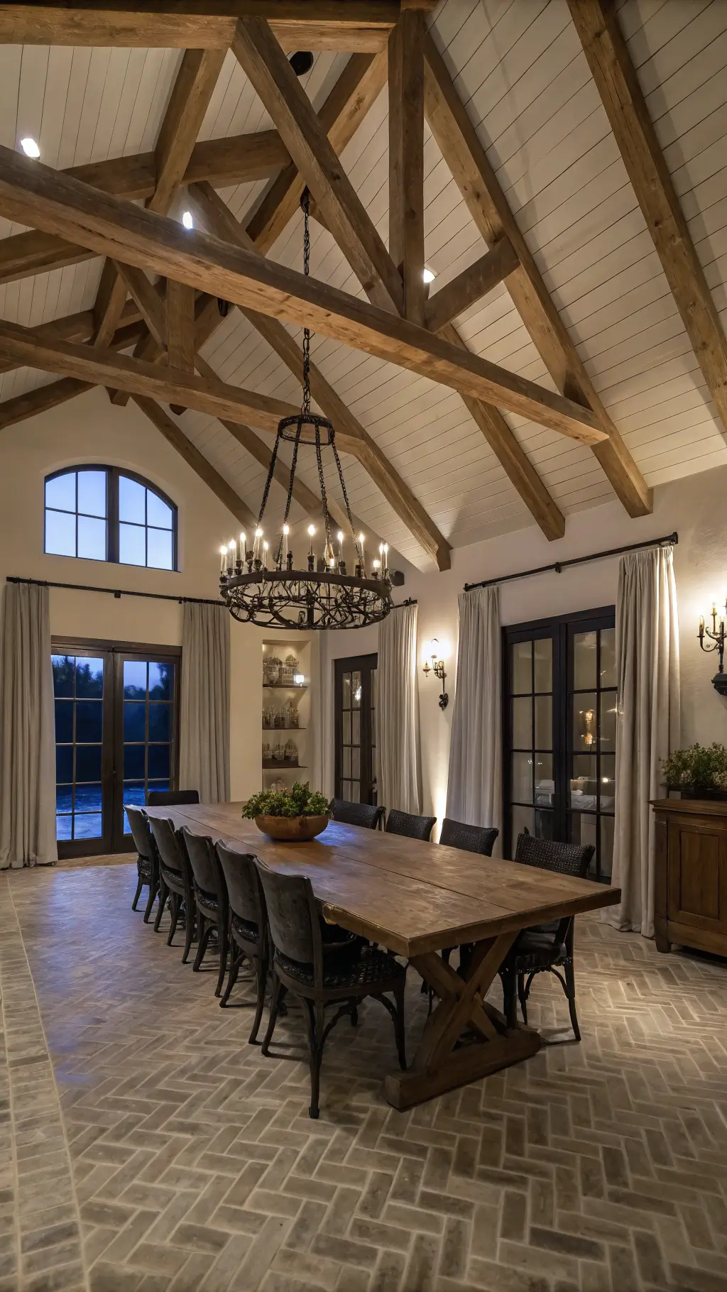 An exquisite cathedral-ceilinged dining room showcasing exposed wooden beams, a grand oak table surrounded by matte black chairs, dramatic chandelier lighting, and elegant detailing with grey linen curtains, an antique dough bowl with preserved boxwood, and black metal lanterns.