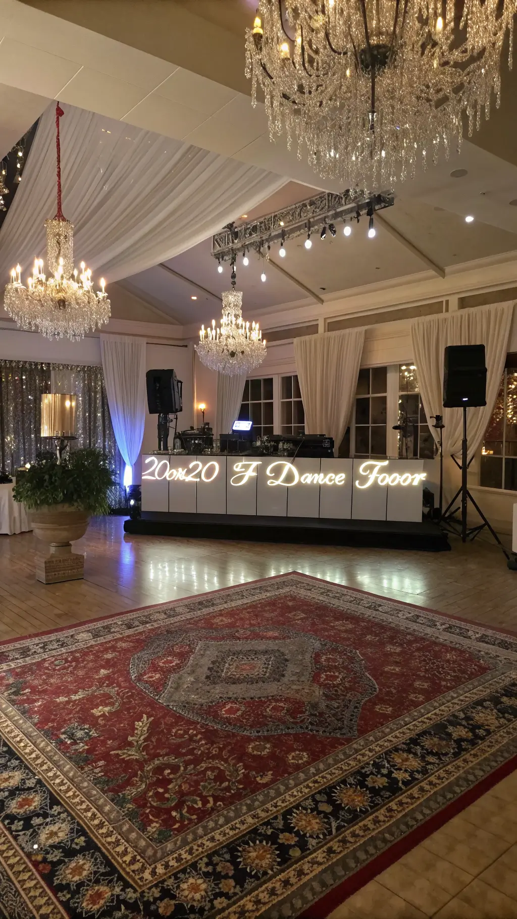 Elevated view of a 20x20ft dance floor layered with vintage Persian rugs and lit by crystal chandeliers and LED light boxes, with metallic fringe curtains behind a modern DJ booth.