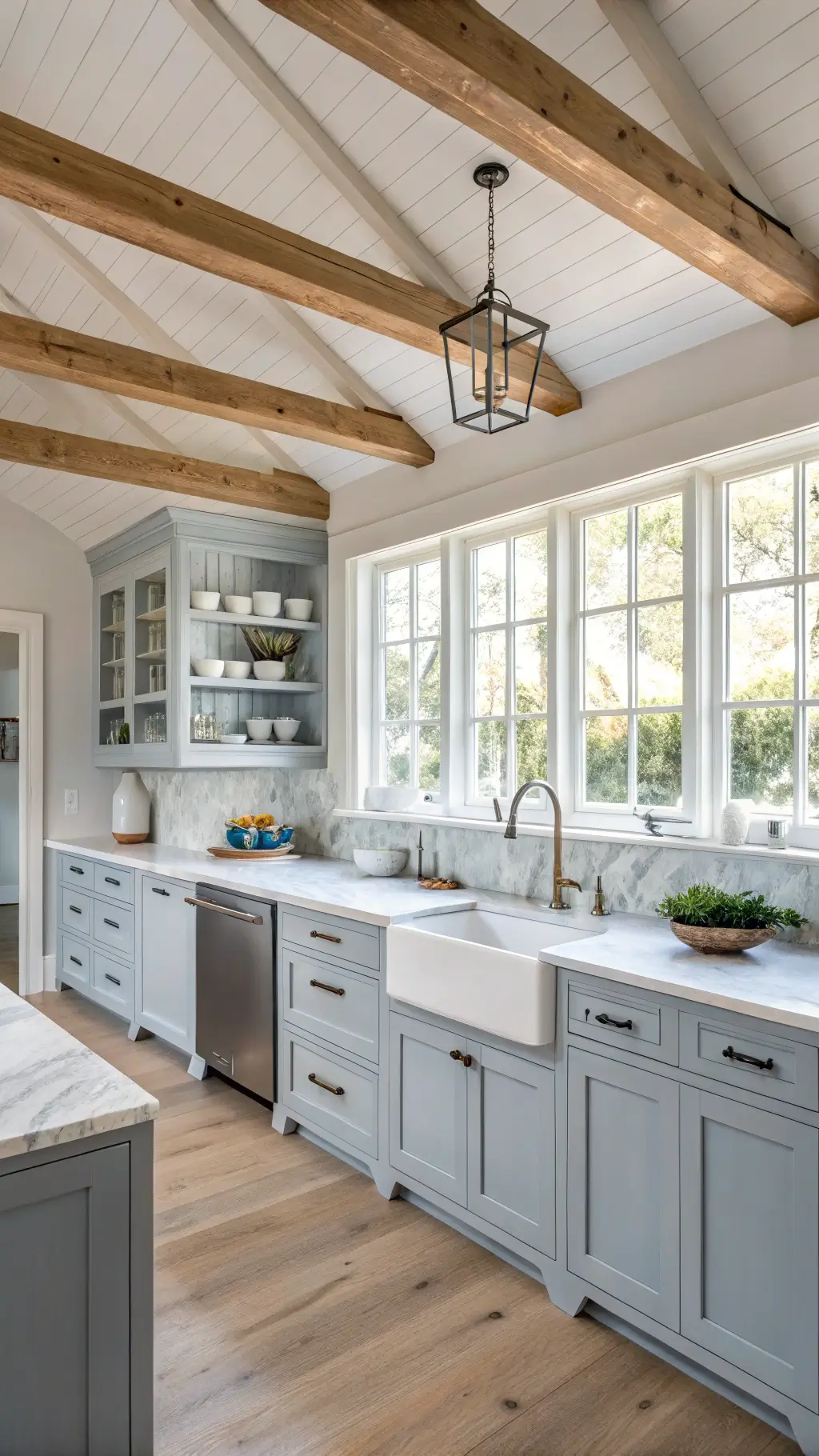 Large, well-lit chef's kitchen with white oak ceiling beams, gray-blue shaker cabinets, honed marble counters, and open shelving featuring white ceramics and sea glass collections, in a refined coastal farmhouse style.